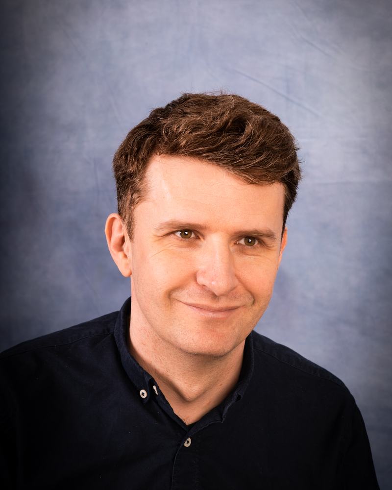 A teacher at IAS Warsaw in a navy shirt, with a gentle smile, studio portrait